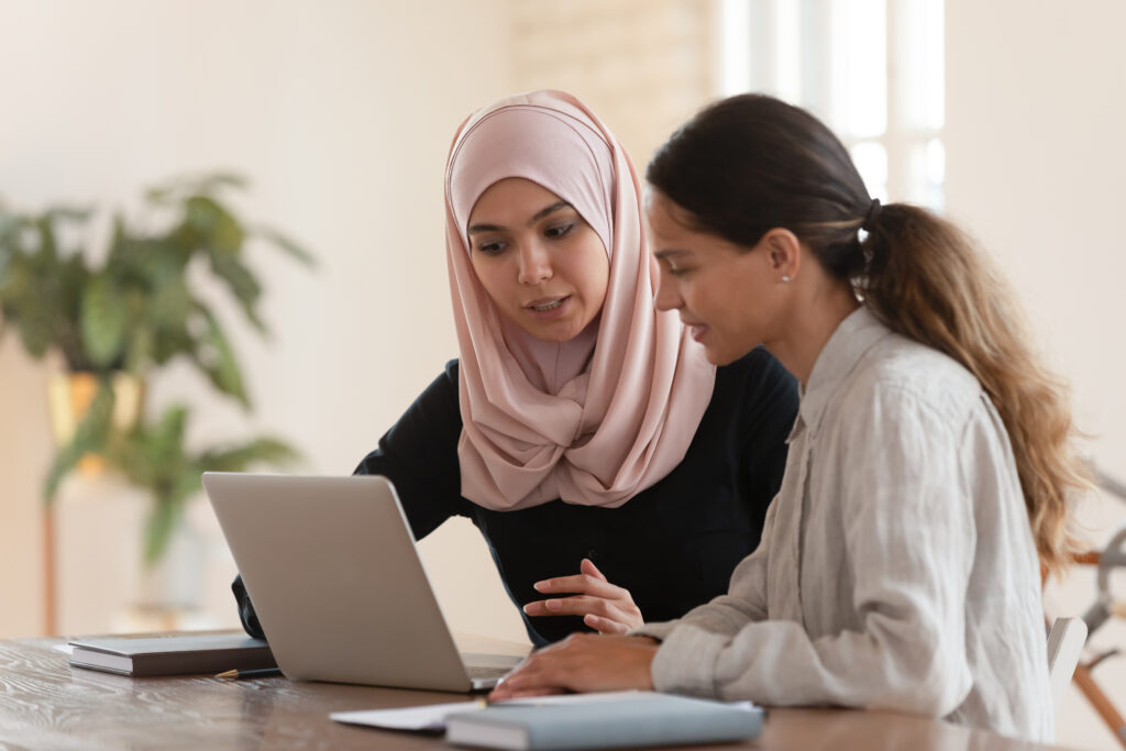 Eine junge Frau mit Kopftuch erklärt einem jungen Mädchen etwas vor einem Laptop sitzend.