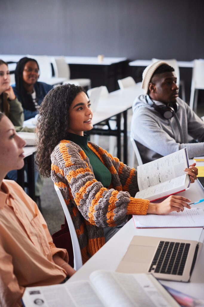 Junge Studentin lächelt während einer Unterrichtsstunde, während sie mit anderen Studenten an einem Schreibtisch in einem Klassenzimmer sitzt.